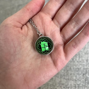 May include: A silver-colored necklace with a round pendant. The pendant features a green gift box design on a black and green background. The necklace is held in a hand.