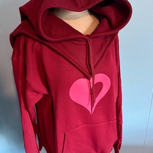 May include: Burgundy hoodie featuring a large pink heart design. The hoodie has a drawstring hood and a kangaroo pocket. The garment is displayed on a mannequin. The background is a light blue wall.