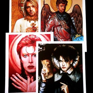 May include: Four different art prints featuring portraits of musicians. The first print features a man with long blonde hair wearing a white shirt and holding a red heart. The second print features a man with long brown hair wearing a red and gold robe with angel wings. The third print features a man with red hair wearing a black shirt and a white halo. The fourth print features a man with black hair wearing a black shirt and holding a small orange cat.