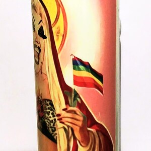 May include: A glass candle jar with a white candle inside. The jar has a colorful image of a person with a rainbow flag. The person is wearing a red robe and a leopard print top.