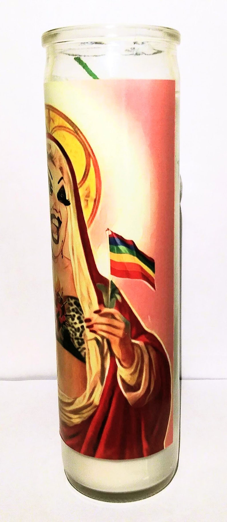May include: A glass candle jar with a white candle inside. The jar has a colorful image of a person with a rainbow flag. The person is wearing a red robe and a leopard print top.