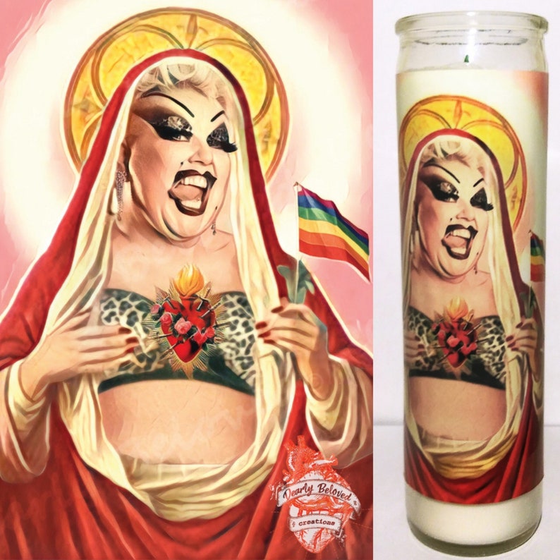 May include: A religious candle featuring a colorful illustration of a woman wearing a red robe and a leopard print top. The woman is holding a rainbow flag and has a heart with flames and roses on her chest. The candle is labeled "Dearly Beloved Creations".