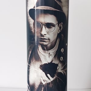 May include: A white glass candle with a black and white image of a man wearing a hat and glasses. The man is holding a small black object in his hand.