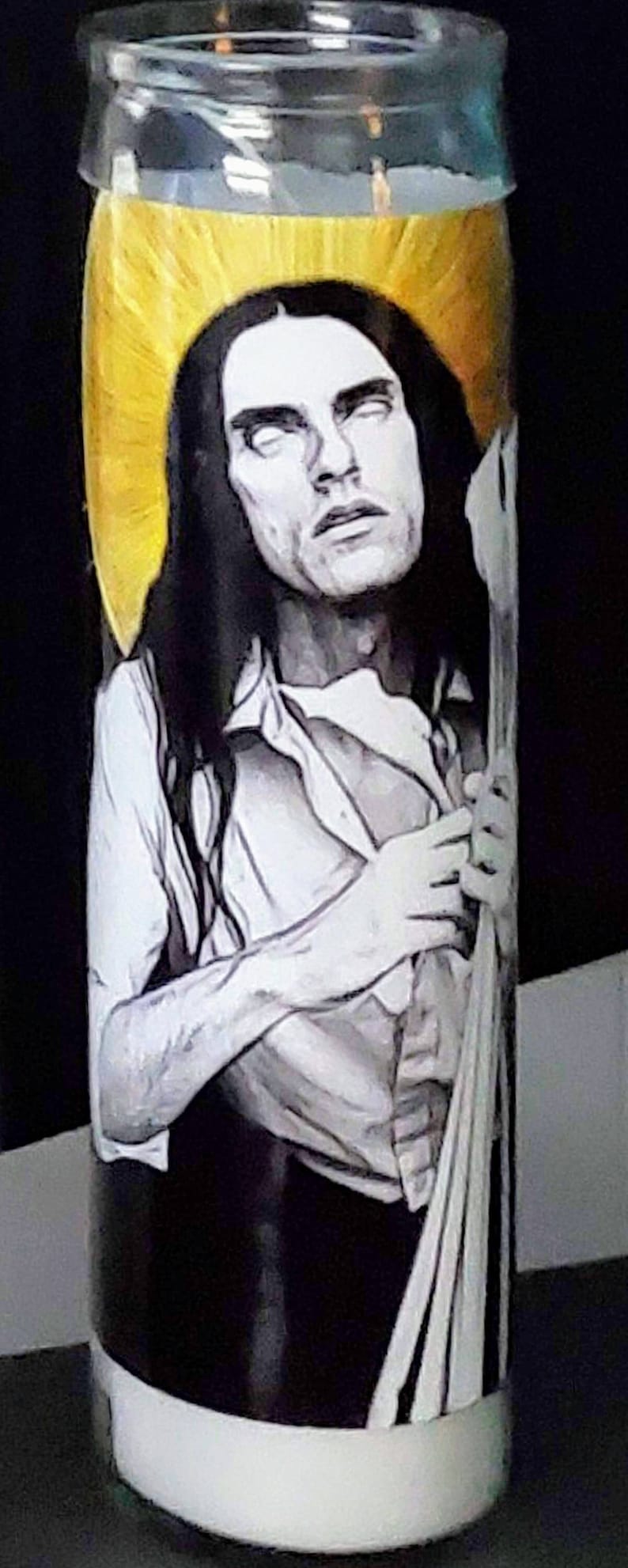 May include: A glass candle jar with a white base and a black and white image of a person with long hair. The person is wearing a white shirt and has a halo of gold light around their head.