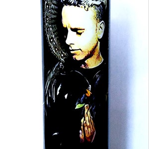 May include: A white glass candle with a black and white image of a person with short, light hair. The person is wearing a black jacket and has their hands clasped in prayer. The image is surrounded by a black and white background.