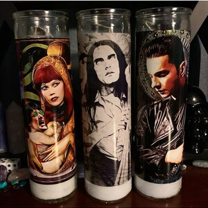 May include: Three glass candle holders with white candles. Each candle holder has a different image printed on it. The first candle holder has a woman with red hair holding a baby. The second candle holder has a man with long black hair. The third candle holder has a man with short black hair.