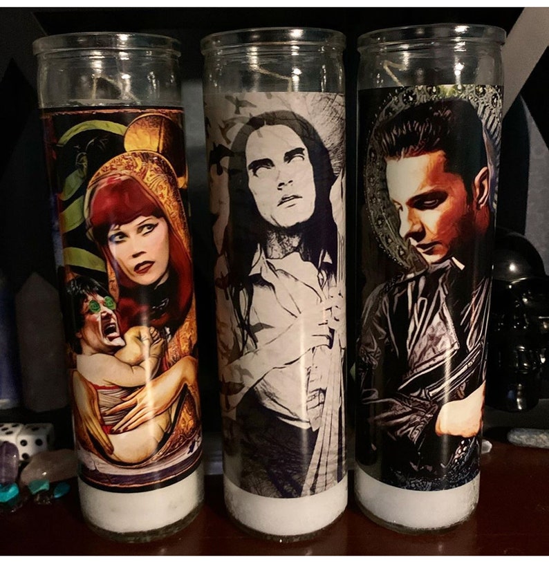 May include: Three glass candle holders with white candles. Each candle holder has a different image printed on it. The first candle holder has a woman with red hair holding a baby. The second candle holder has a man with long black hair. The third candle holder has a man with short black hair.