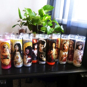 May include: A collection of 12 colorful candles with images of various people on them. The candles are arranged in a row on a dark wooden surface.