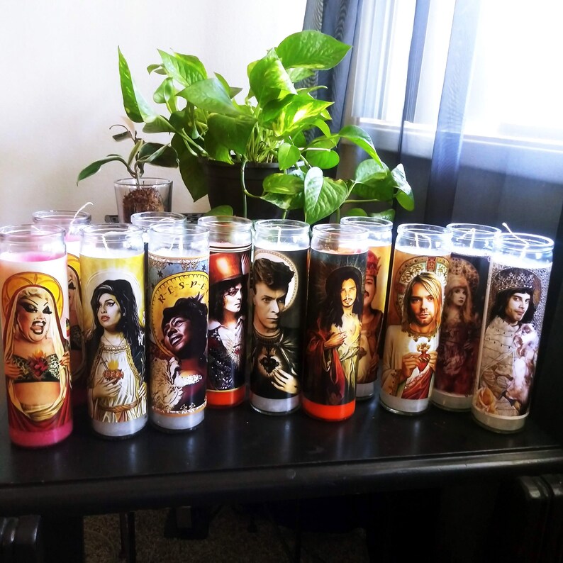 May include: A collection of 12 colorful candles with images of various people on them. The candles are arranged in a row on a dark wooden surface.