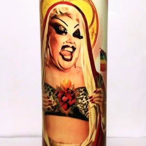 May include: A glass candle with a colorful image of a person wearing a red and gold robe and a leopard print top. The person has a heart with flames on their chest and is holding a rainbow flag.