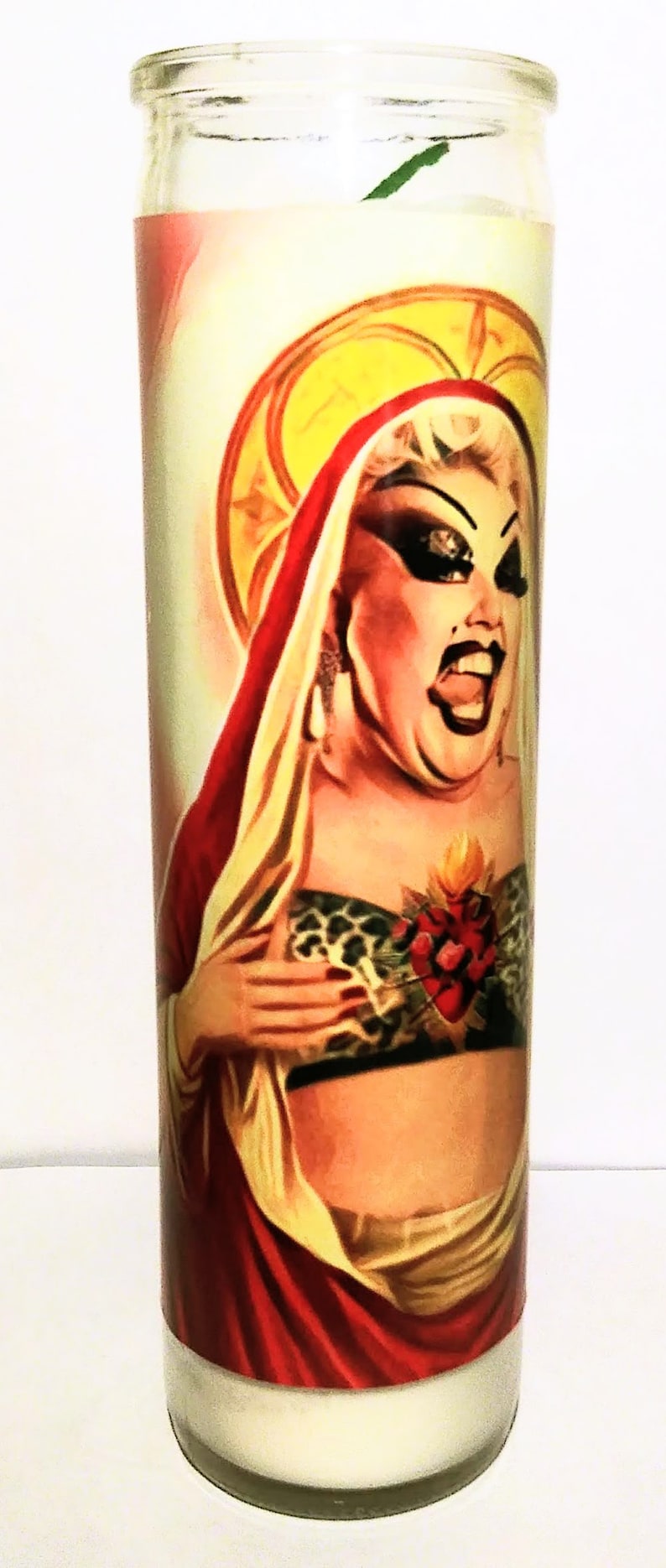 May include: A white candle with a colorful image of a person wearing a red robe and a gold crown. The person has a large heart with flames on their chest. The image is surrounded by a yellow halo.