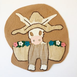 May include: A felt applique of a white cow wearing a brown hat and overalls, holding a basket of flowers. The flowers are pink, white, and blue.
