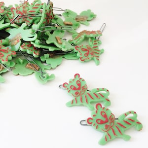 May include: A pile of green plastic tiger hair clips with red stripes and red eyes. There are two clips in the foreground, one above the other.