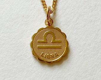 May include: Gold necklace with a Libra zodiac sign pendant. The pendant is round with a scalloped edge and has a textured surface. The Libra symbol is raised and centered on the pendant.