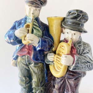 May include: A vintage ceramic figurine of two musicians playing instruments. The musician on the left is wearing a blue jacket and playing a trumpet. The musician on the right is wearing a gray jacket and playing a tuba. Both figurines are painted in a colorful, glazed style.