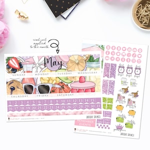 May include: A purple and pink monthly calendar planner with the word "May" in a large font. The calendar features a variety of illustrations, including a strawberry, a glass of juice, a pair of sunglasses, and a cake. The calendar is decorated with glitter and foil accents.