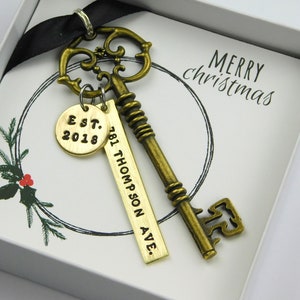 May include: An antique brass key with a decorative design, paired with a gold tag engraved with "Est. 2018" and a rectangular tag with an address. The key and tags are presented in a gift box with a Christmas greeting.