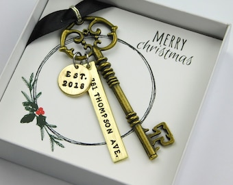 Puede incluir: Una llave antigua de latón con un diseño decorativo, combinada con una etiqueta dorada grabada con "Est. 2018" y una etiqueta rectangular con una dirección. La llave y las etiquetas se presentan en una caja de regalo con un saludo navideño.