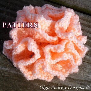 May include: A close-up of a peach-colored crocheted flower with a ruffled texture. The flower is on a wooden surface. The word "PATTERN" is written in red letters above the flower.