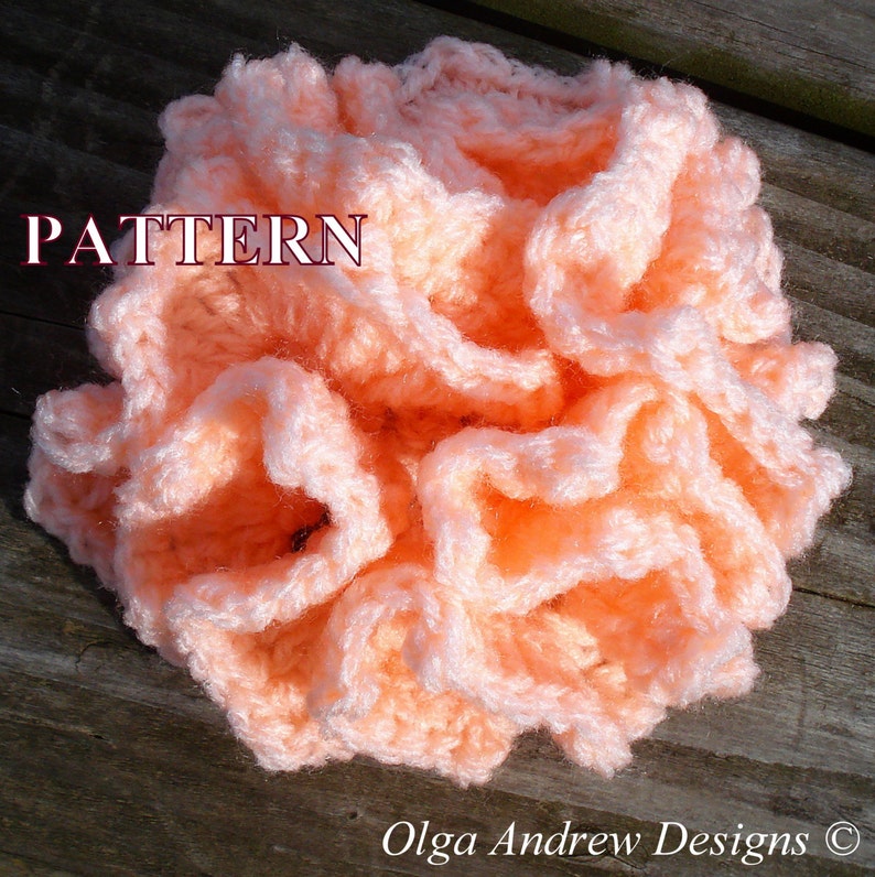 May include: A close-up of a peach-colored crocheted flower with a ruffled texture. The flower is on a wooden surface. The word "PATTERN" is written in red letters above the flower.