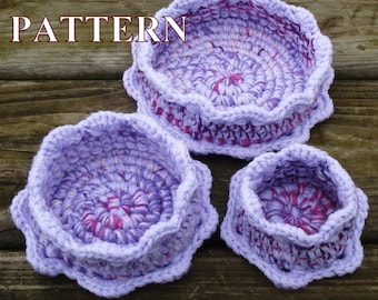 Puede incluir: Tres cestas de crochet en tonos de morado y rosa. Las cestas están anidadas una dentro de la otra, con la cesta más grande en la parte superior y la cesta más pequeña en la parte inferior. Las cestas tienen un borde festoneado y un diseño texturizado.