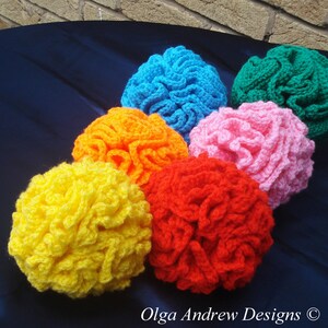 K&ouml;nnte beinhalten: Eine Sammlung von sechs geh&auml;kelten Gegenst&auml;nden in leuchtenden Farben: Gelb, Orange, Rot, Rosa, Blau und Gr&uuml;n. Die Artikel haben ein strukturiertes, ger&uuml;schtes Aussehen. Das Logo von Olga Andrew Designs ist unten rechts sichtbar.
