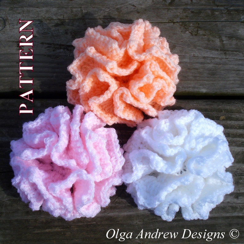 May include: Three crocheted flowers in pink, white, and peach colors. The flowers are made with a ruffled stitch and are arranged on a wooden surface.