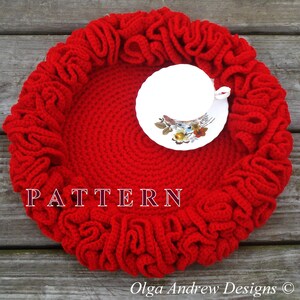 May include: A bright red, crocheted tea cup coaster with a ruffled edge. A white teacup and saucer with a floral design sit in the center. The word "PATTERN" is displayed in white block letters.