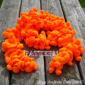 May include: A bright orange crocheted scarf with a textured, puffy design. The scarf is laid out on a wooden surface. The text "PATTERN" is visible in the center of the image. The text "Olga Andrew Designs ©" is visible at the bottom of the image.