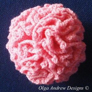 K&ouml;nnte beinhalten: Eine handgefertigte, runde, rosa H&auml;kelblume. Die Blume besteht aus Garn und hat ein strukturiertes, ger&uuml;schtes Aussehen. Der Text "Olga Andrew Designs" befindet sich unten rechts.