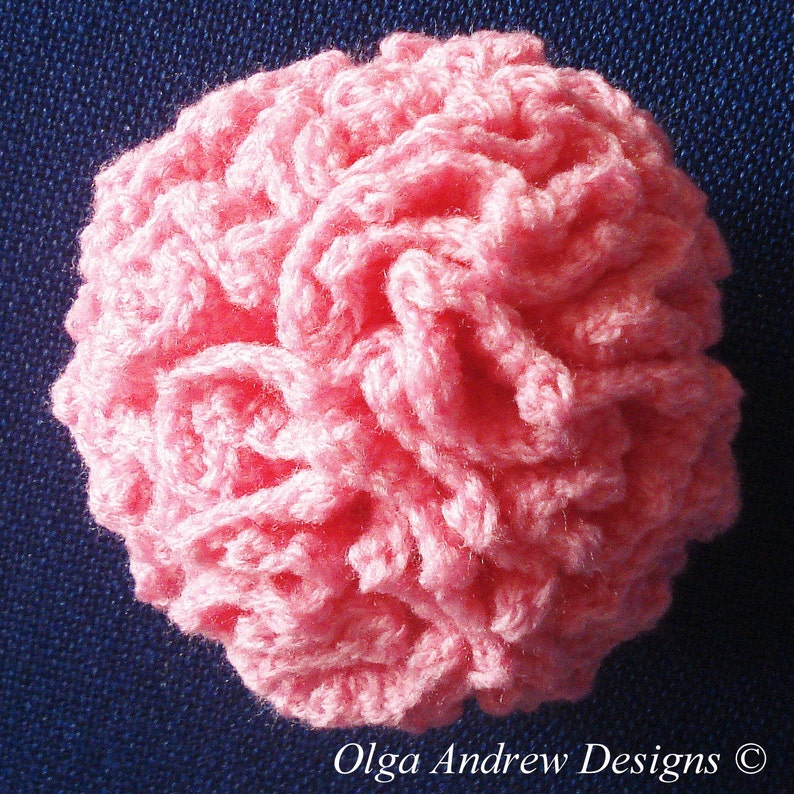 K&ouml;nnte beinhalten: Eine handgefertigte, runde, rosa H&auml;kelblume. Die Blume besteht aus Garn und hat ein strukturiertes, ger&uuml;schtes Aussehen. Der Text "Olga Andrew Designs" befindet sich unten rechts.