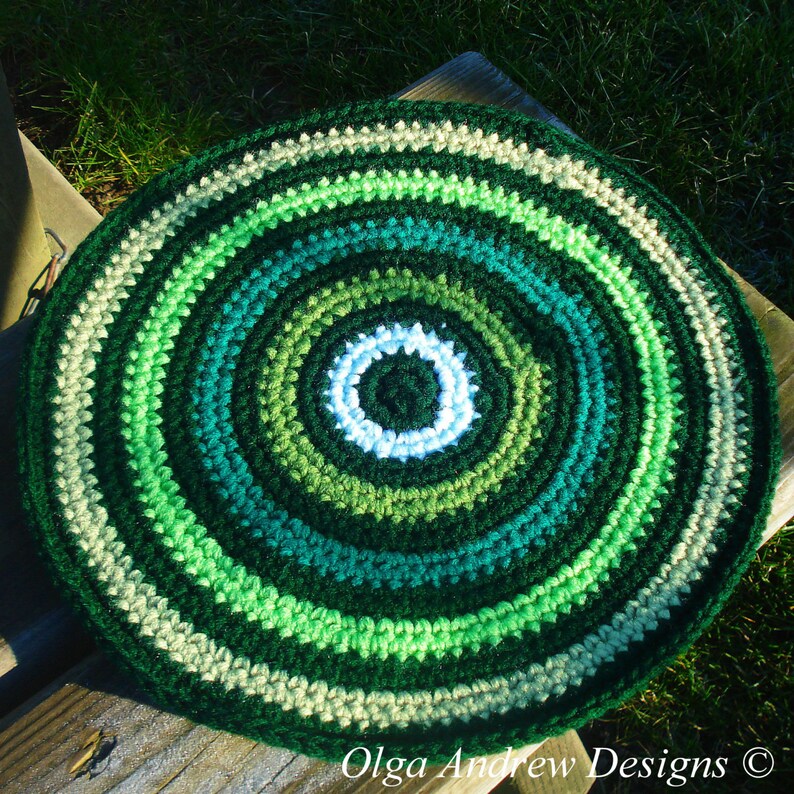 May include: A round crocheted rug with a concentric circle pattern in shades of green, white, and beige. The rug has a center circle with a white ring and a dark green center.