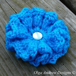May include: A bright blue crocheted flower with a white button in the center. The flower is handmade and has a textured, ruffled appearance.