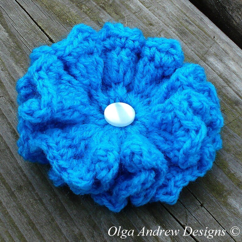 May include: A bright blue crocheted flower with a white button in the center. The flower is handmade and has a textured, ruffled appearance.