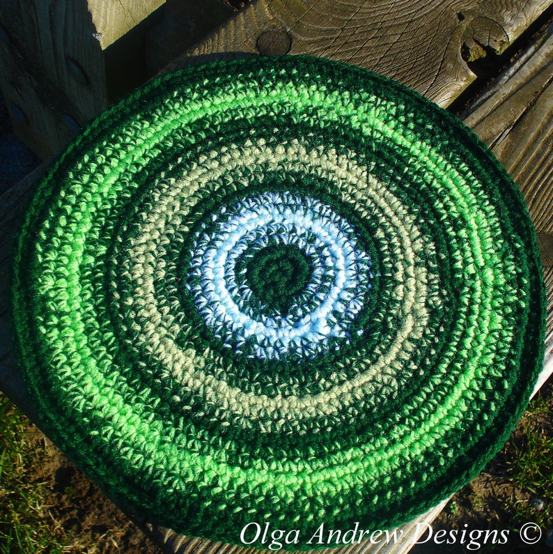 May include: A green, white, and beige crocheted round rug with a concentric circle pattern. The rug is made with a variety of shades of green, white, and beige yarn.