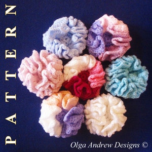 May include: A collection of seven crocheted flowers in various colors, including pink, blue, white, and purple. The flowers are arranged in a circle on a blue background. The text "Olga Andrew Designs &copy;" is visible at the bottom of the image.
