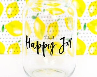 Może przedstawiać: Szklany słoik z przezroczystego szkła z białą pokrywką. Słoik ma czarną etykietę z napisem "The Happy Jar".