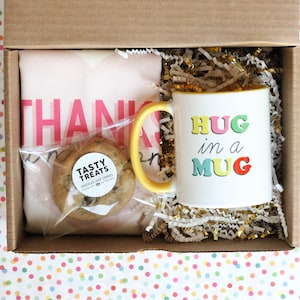 May include: A gift box filled with a pink tote bag with the words "Thank You" printed on it, a yellow mug with the text "Hug in a Mug" in colourful letters, and a chocolate chip cookie in a clear plastic bag.