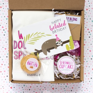 May include: A gift box containing a white t-shirt with pink text, a greeting card with a sloth illustration, a cookie, a candle, and a necklace. The card reads "Happy belated Birthday". The candle is labeled "Sprinkled Cupcake".