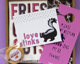 May include: A gift box filled with a variety of items, including a t-shirt with the text "FRIES love stinks GUY", a chocolate chip cookie, a card with a skunk illustration and the text "love stinks", and a pink door hanger with the text "PLEASE NOT DISTURB 000-ING MY EX. THANKS."