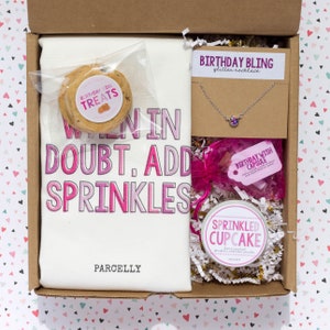 May include: A gift box filled with birthday themed items, including a white t-shirt with the text "When in doubt, add sprinkles", a cookie, a necklace, a candle, and a small jar of scented candle.