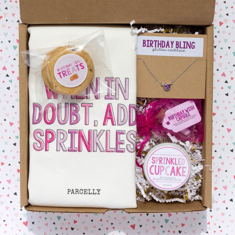 May include: A gift box filled with birthday themed items, including a white t-shirt with the text "When in doubt, add sprinkles", a cookie, a necklace, a candle, and a small jar of scented candle.