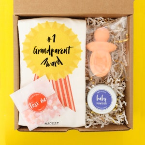 May include: A brown cardboard gift box with a white and red striped tote bag, a pink baby doll, a small jar of baby powder scented candle, and a bag of confetti. The tote bag has a yellow star with the text "#1 Grandparent Award".