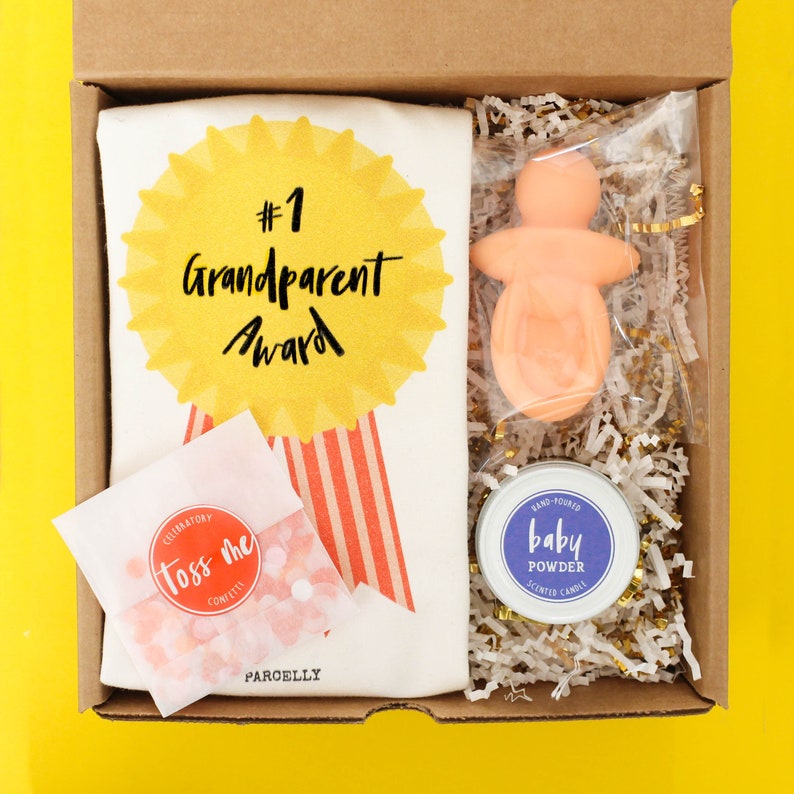 May include: A brown cardboard gift box with a white and red striped tote bag, a pink baby doll, a small jar of baby powder scented candle, and a bag of confetti. The tote bag has a yellow star with the text "#1 Grandparent Award".