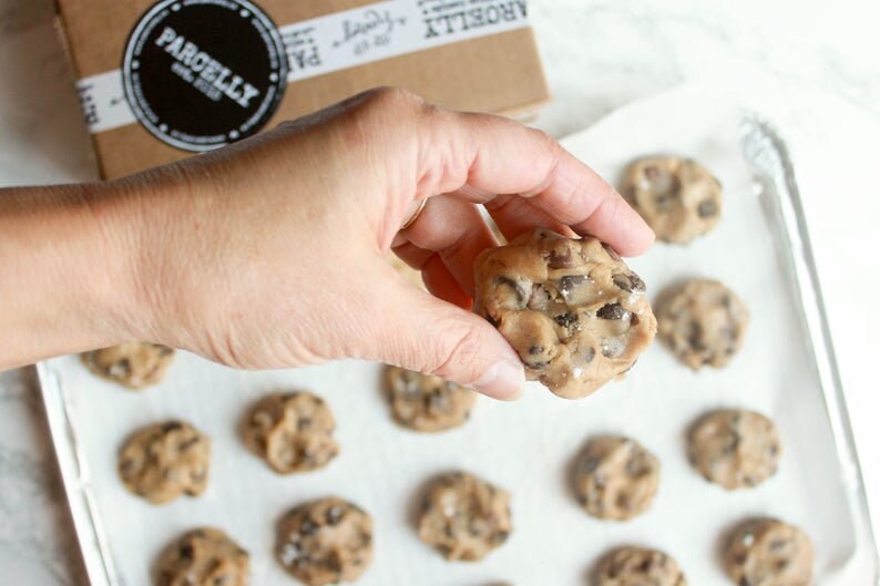 May include: A hand holding a raw chocolate chip cookie dough ball, with other cookie dough balls on a baking sheet. The dough is speckled with chocolate chips and sea salt. A box with the text "PARCELLY" is in the background.