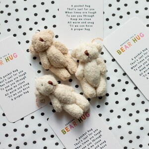 May include: Three small, cream-coloured teddy bears are arranged on a white background with black polka dots. Each bear is holding a card with the text "Sending you a Bear Hug" and a poem about a pocket hug.