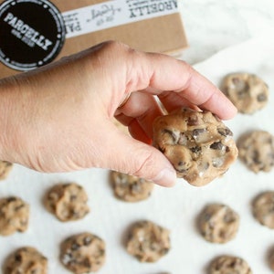 May include: A hand holds a raw chocolate chip cookie dough ball over a baking sheet lined with parchment paper. The cookie dough is surrounded by other cookie dough balls, all ready to be baked.