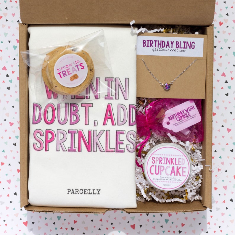 May include: A gift box filled with a white tea towel with the text "When in doubt, add sprinkles", a cookie, a silver necklace with a purple gem, a candle, and a "Birthday Wish Capsule". The box is filled with decorative paper.