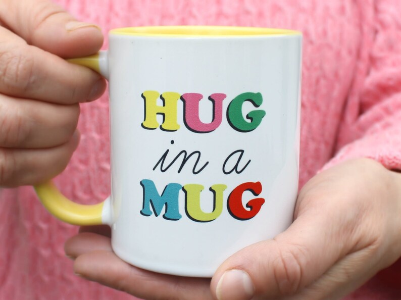 May include: White ceramic mug with a yellow rim and handle. The mug has the text "HUG in a MUG" printed on it in colourful letters.