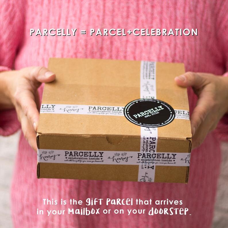 May include: A brown cardboard box with white tape that says "PARCELLY" and "Hip Hip Hooray!"  The box is being held by a person wearing a pink sweater. The text on the box reads "PARCELLY = PARCEL + CELEBRATION".  This is the gift parcel that arrives in your mailbox or on your doorstep.